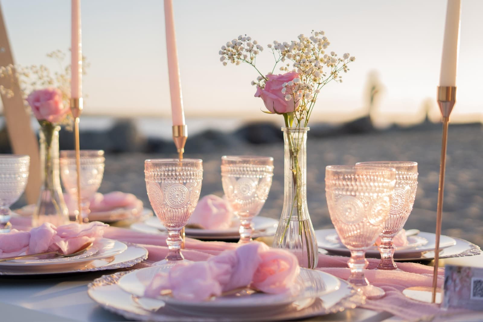 A sunset beach picnic place setting with pink pressed-glass goblets, blush napkins, and bud vases of pink roses and baby’s-breath