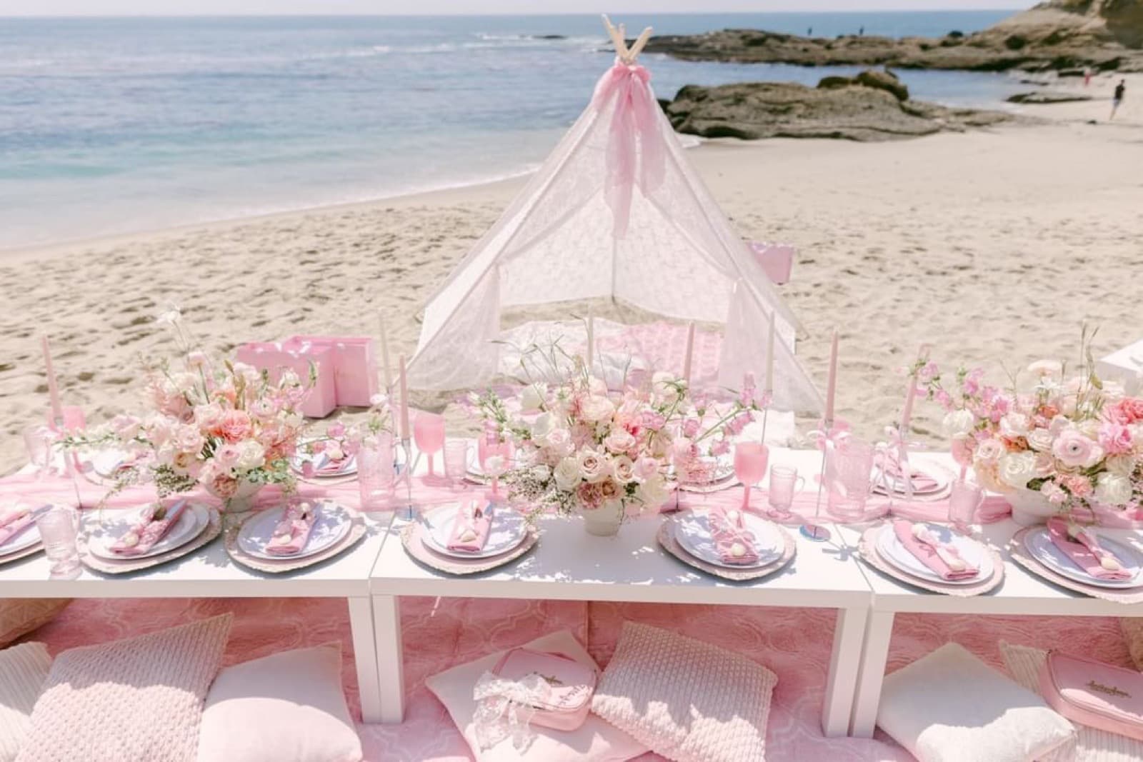 A daytime pink beach picnic with a lace teepee and multiple floral centerpieces, the ocean behind