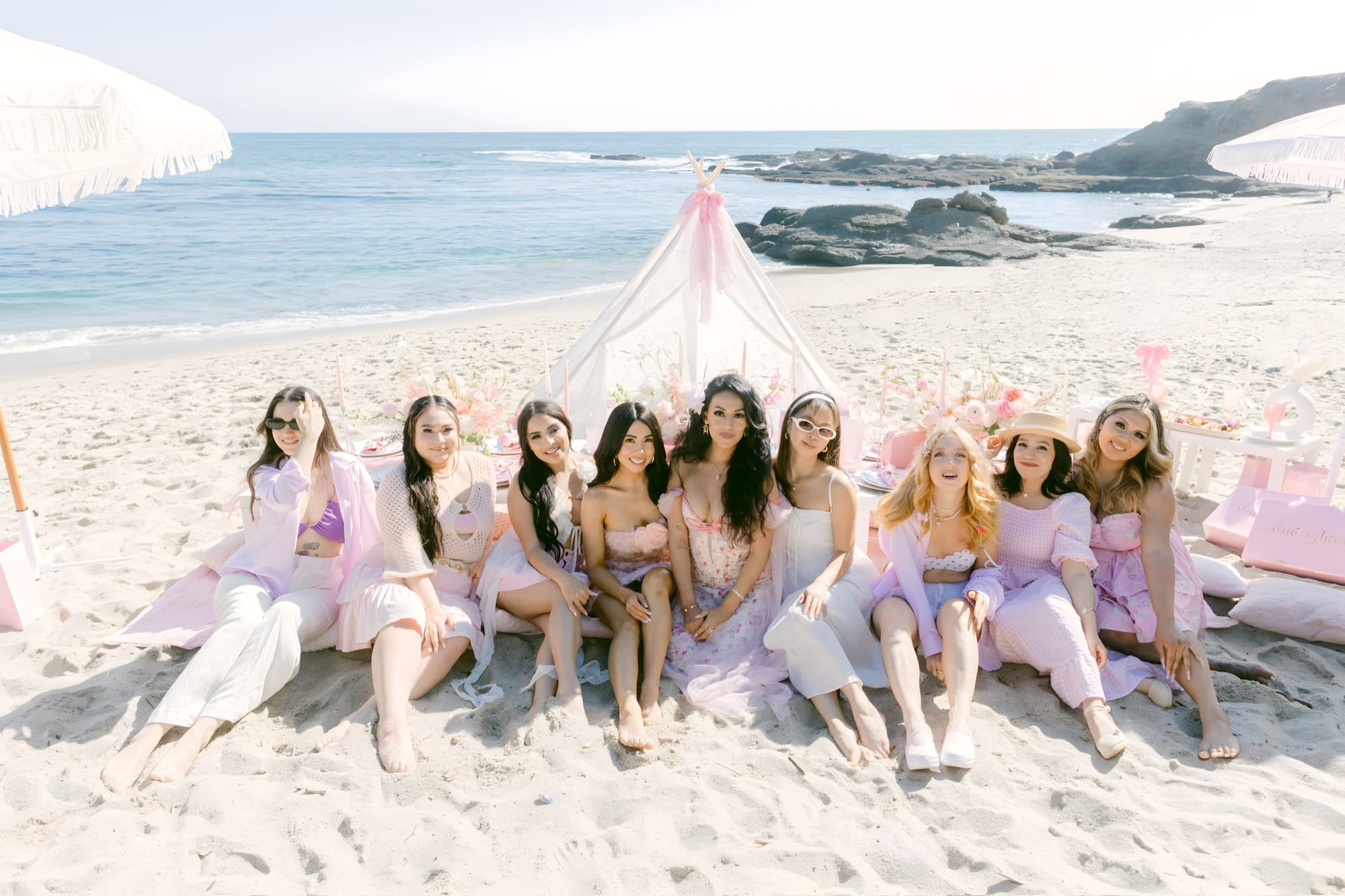 A group of guests in soft pinks gathered on the sand before a lace teepee at a styled Pixxie beach picnic, the ocean behind at golden hour
