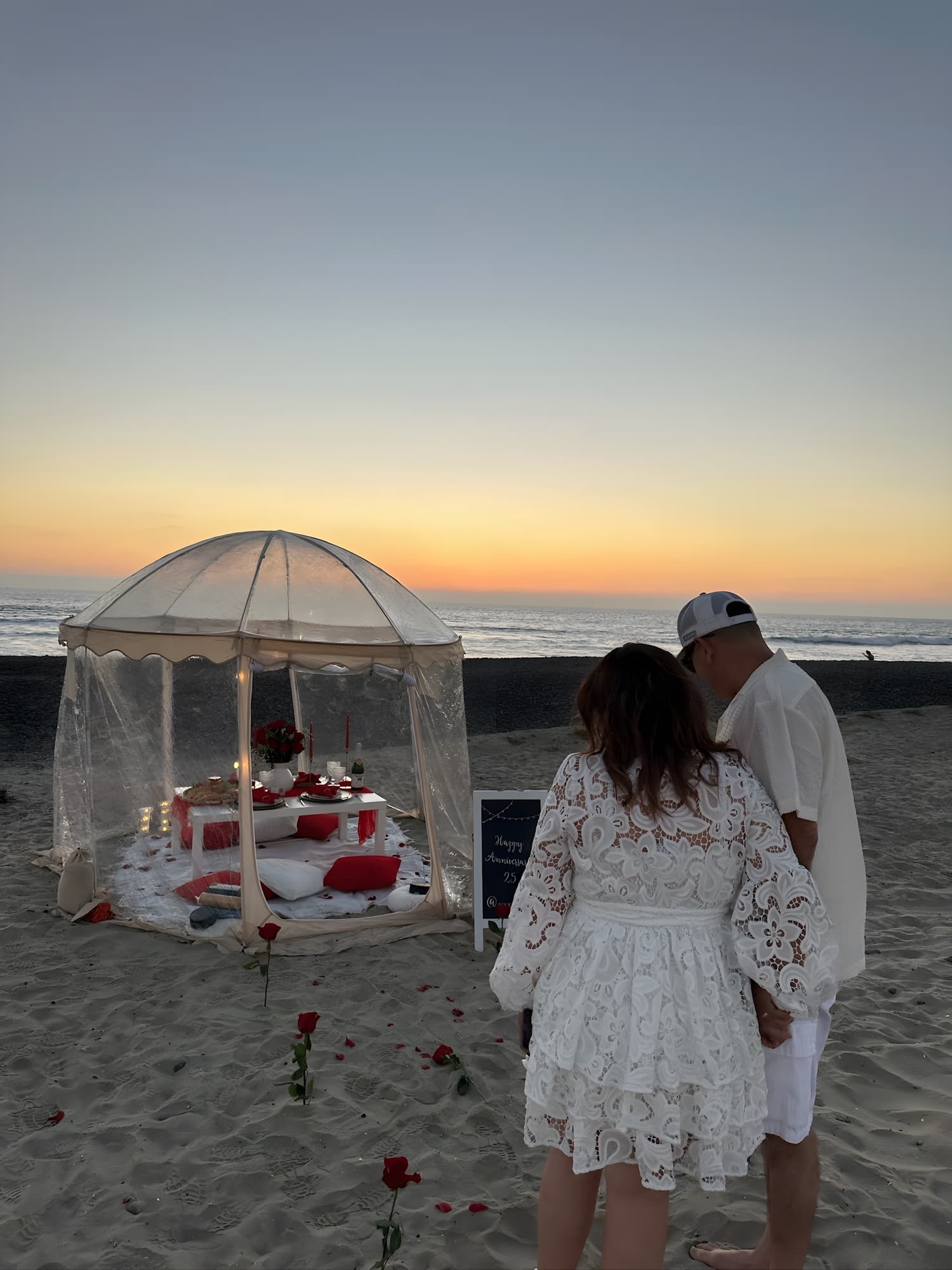 A couple embracing at golden hour beside a private clear-dome beach picnic set with candles and roses, the sunset ocean behind