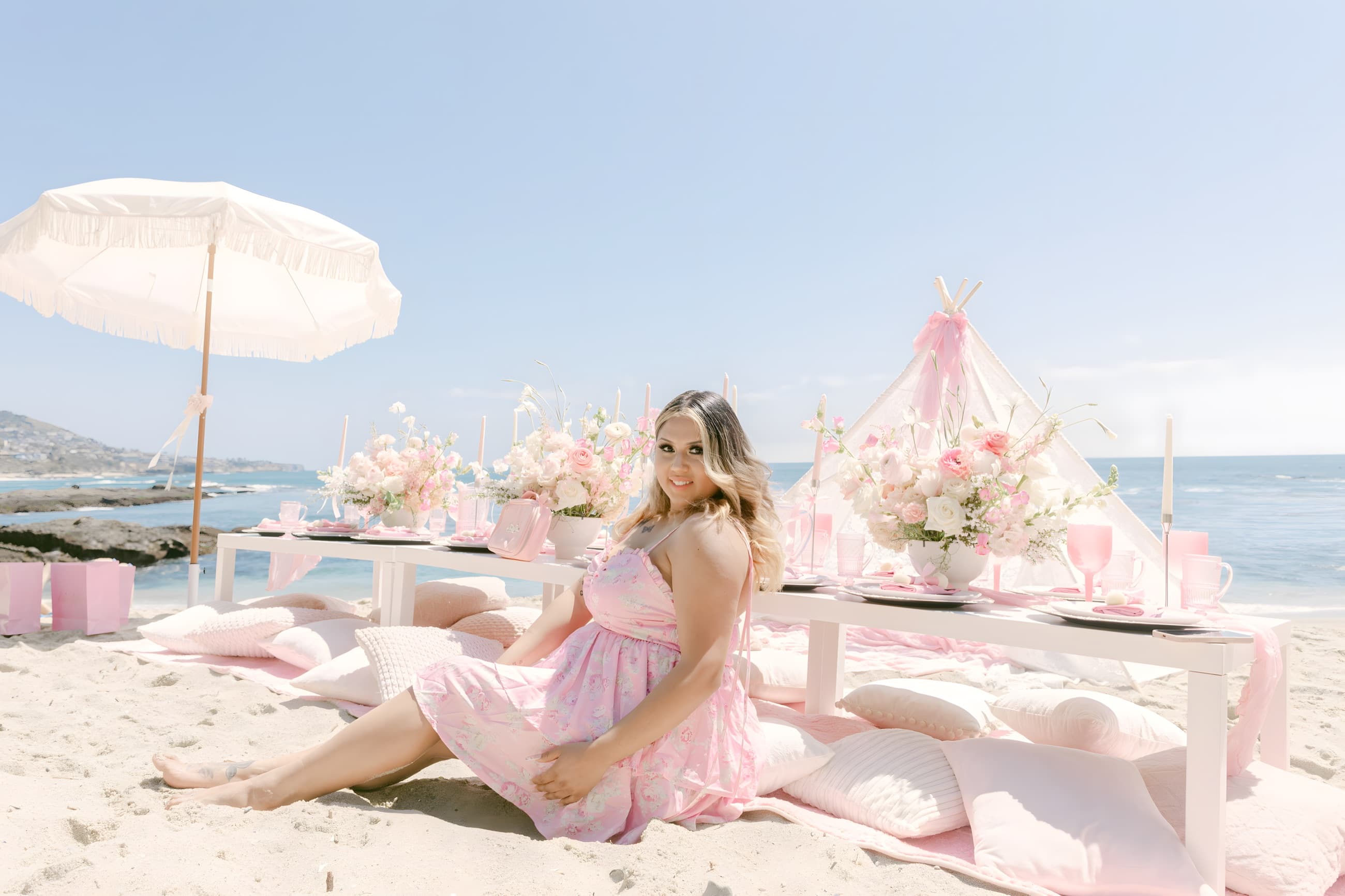 Gina, the founder of Pixxie Picnics, in a pink floral dress seated on the sand and smiling, her full styled pink beach picnic behind her