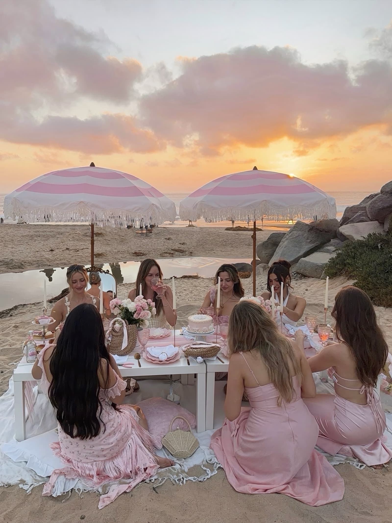 A group of women in soft pinks gathered around a styled blush beach picnic table at sunset, beneath two pink-striped fringed umbrellas, the ocean behind