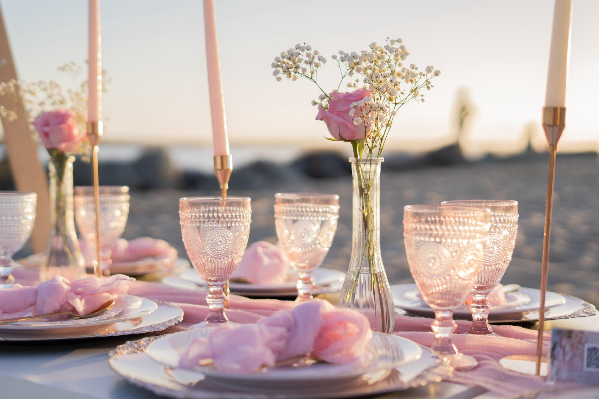 A golden-hour blush beach place setting with pink pressed-glass goblets, blush gauze napkins, rose-gold tapers and a bud vase of a pink rose and baby’s-breath
