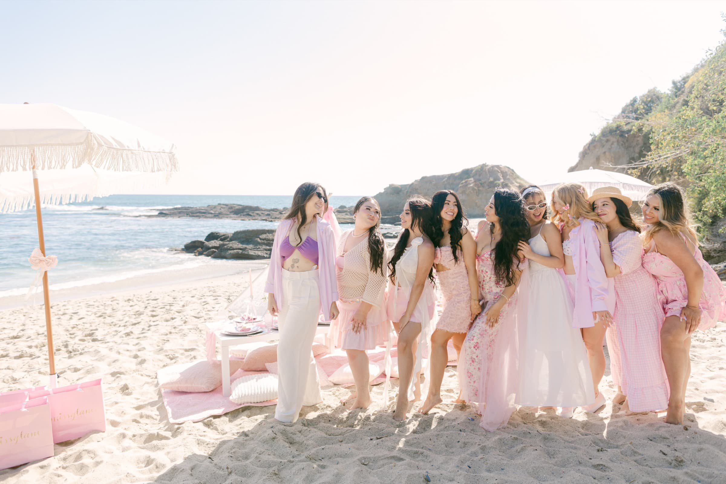A group of guests in soft pinks gathered on a sunlit beach beside a styled Pixxie picnic with a fringed umbrella and floor cushions