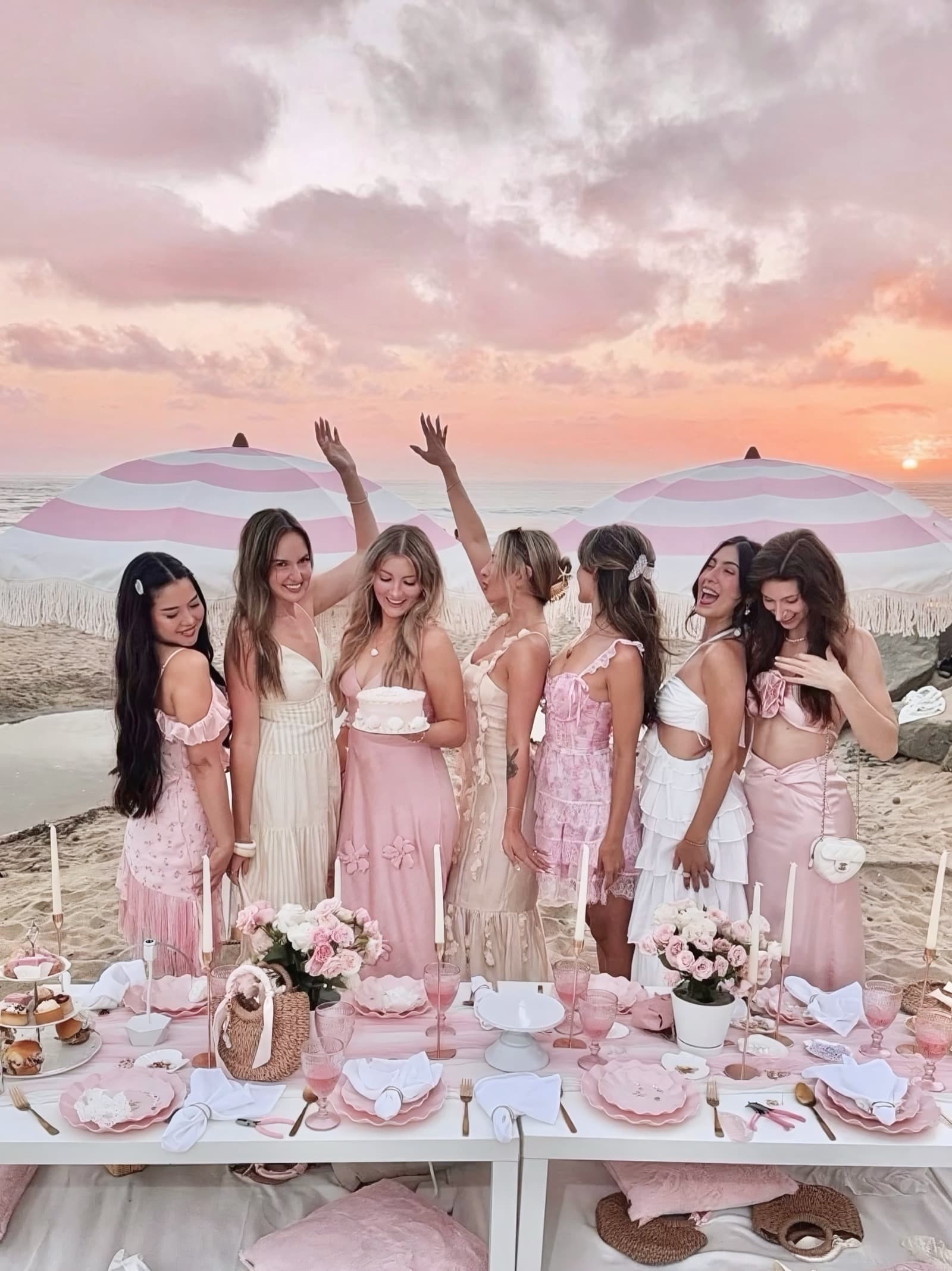 Seven women in soft pinks and cream standing together behind a styled blush beach picnic table at sunset