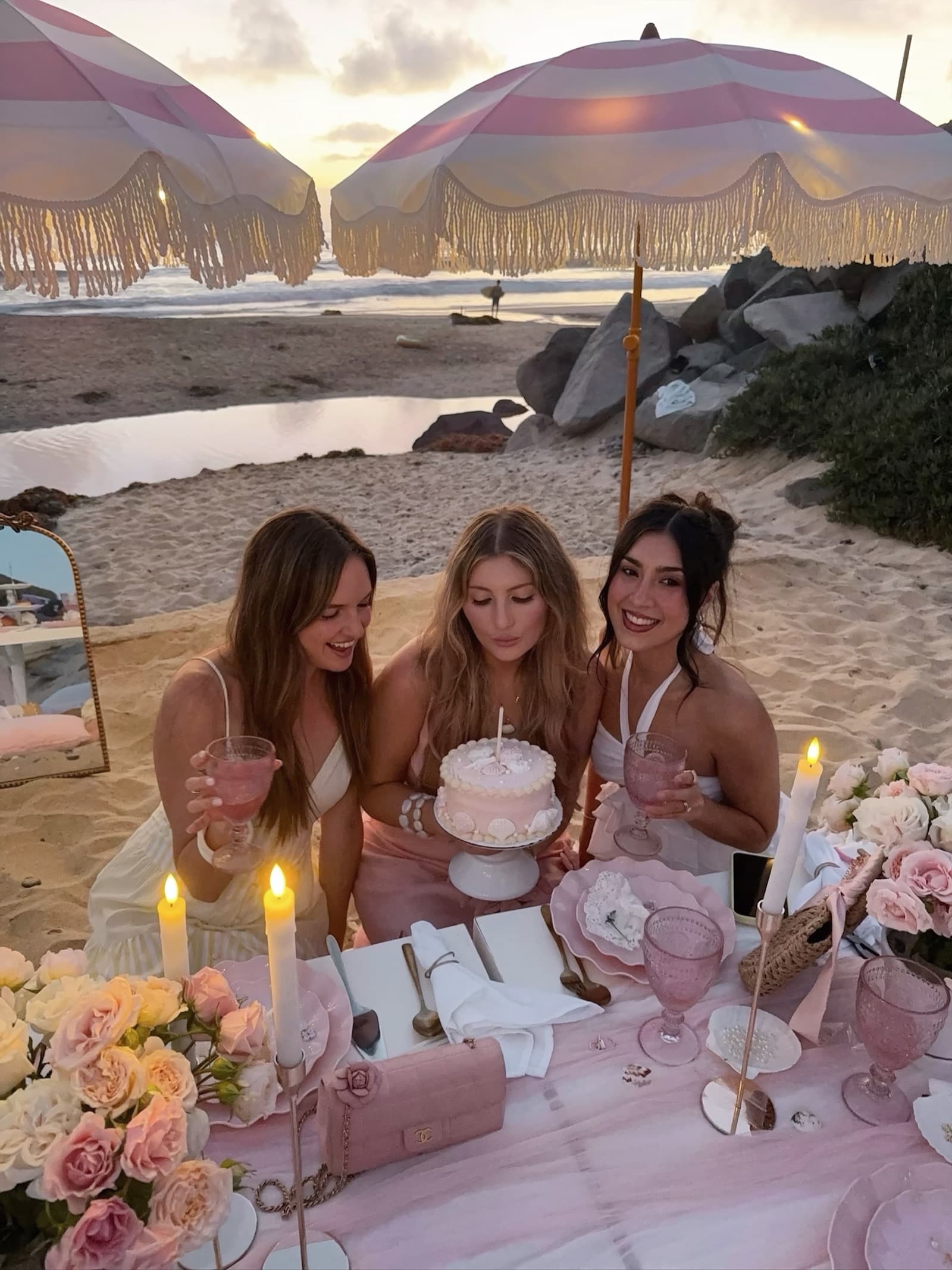 Three women in soft pinks laughing around a small shell-decorated cake at a blush beach picnic in warm sunset light