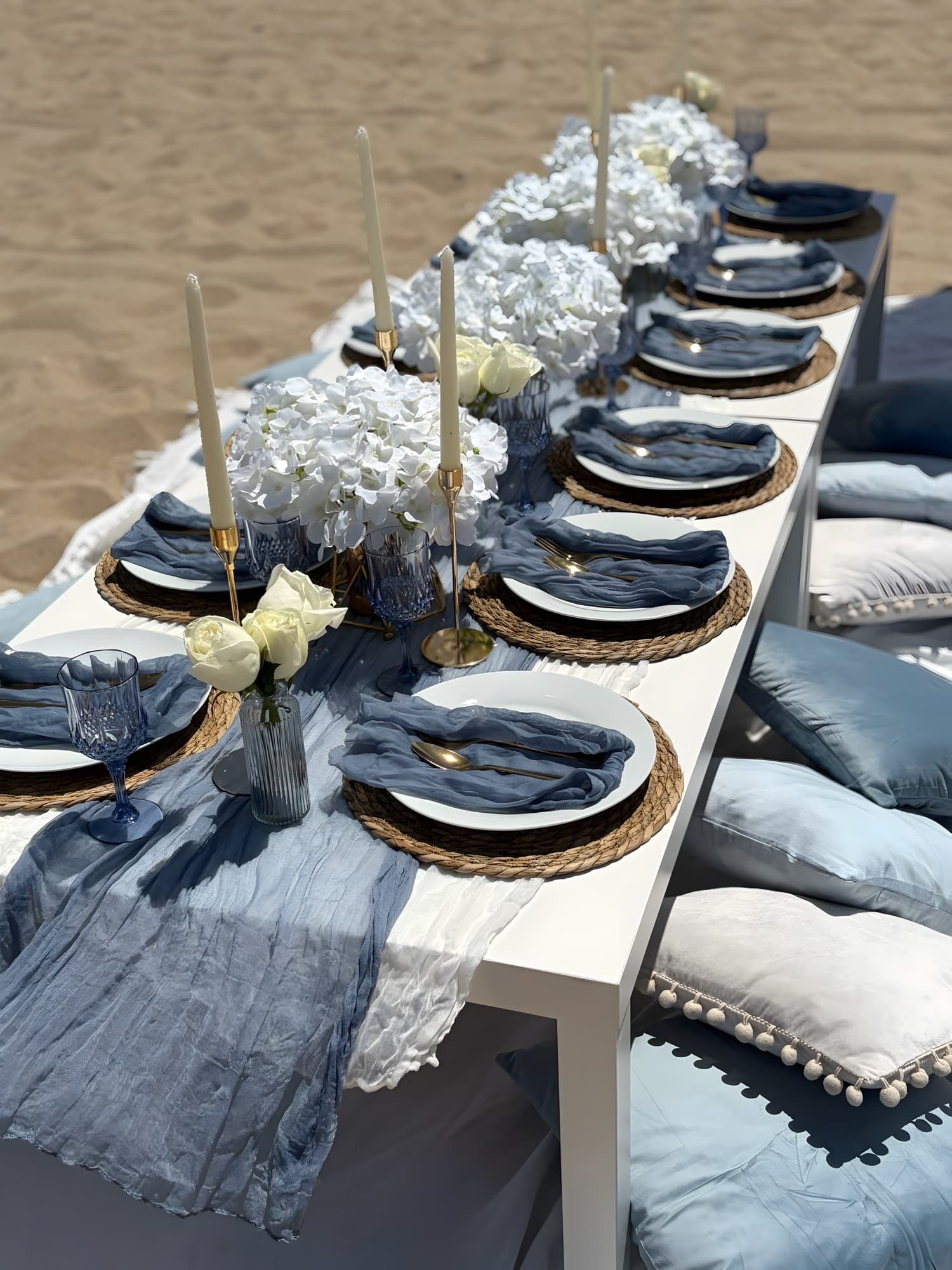 A dusty-blue beach tablescape with blue gauze runners, white hydrangea, gold flatware and blue glassware on the sand