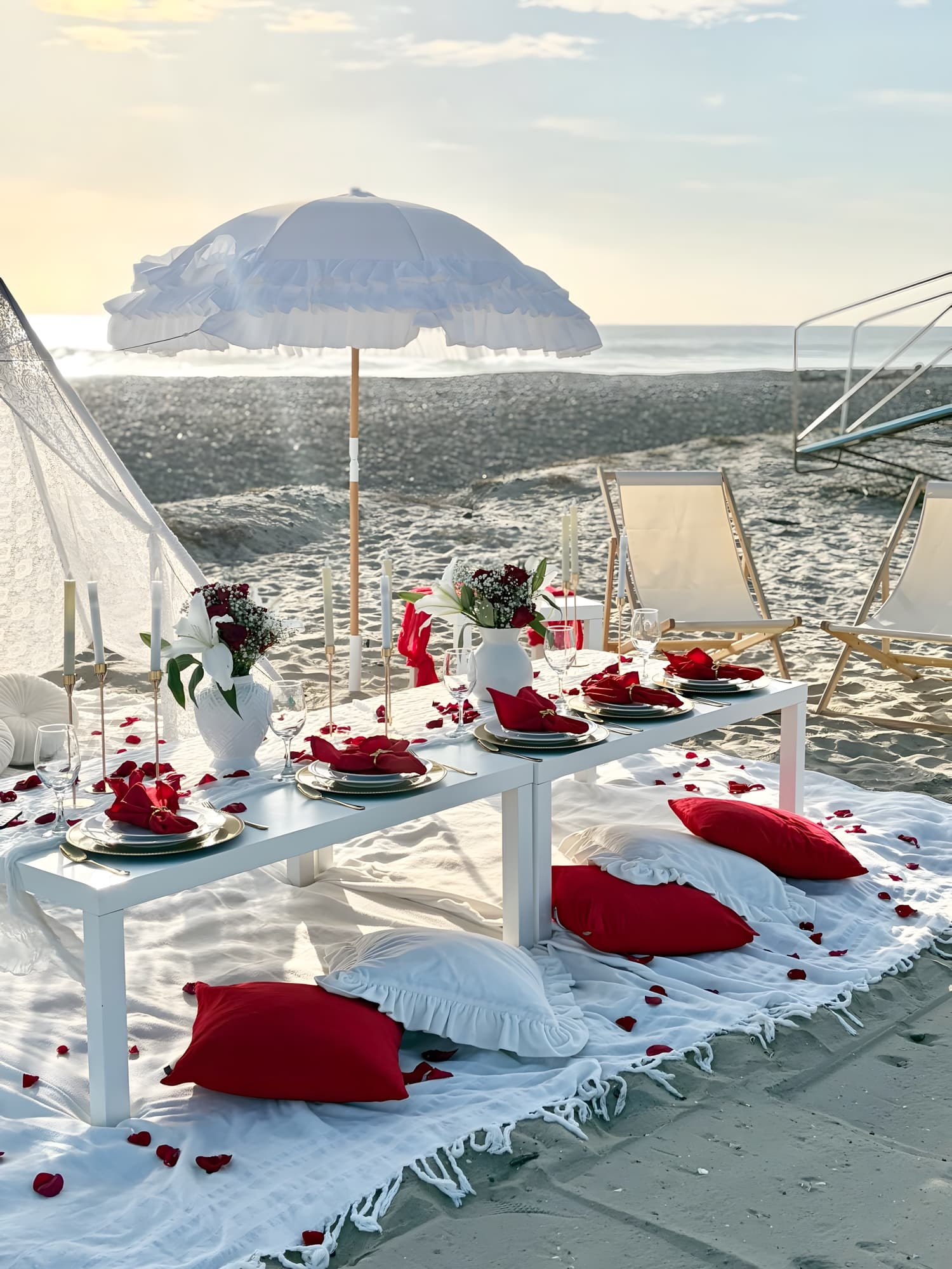 A romantic beach picnic with a white ruffled umbrella, red roses, red napkins and rose petals at golden hour