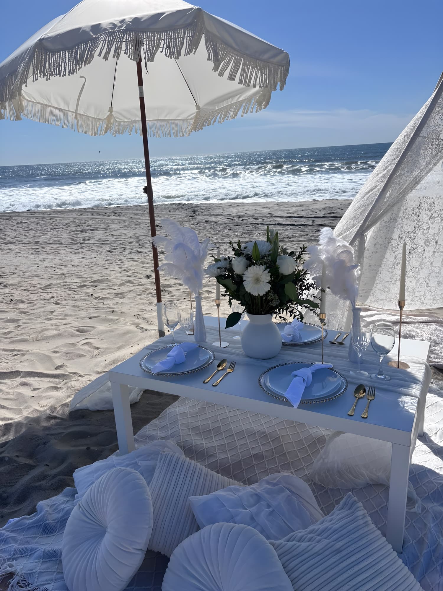 An ivory beachfront picnic with white ostrich-feather accents, a fringed umbrella and a white floral centerpiece, the ocean behind