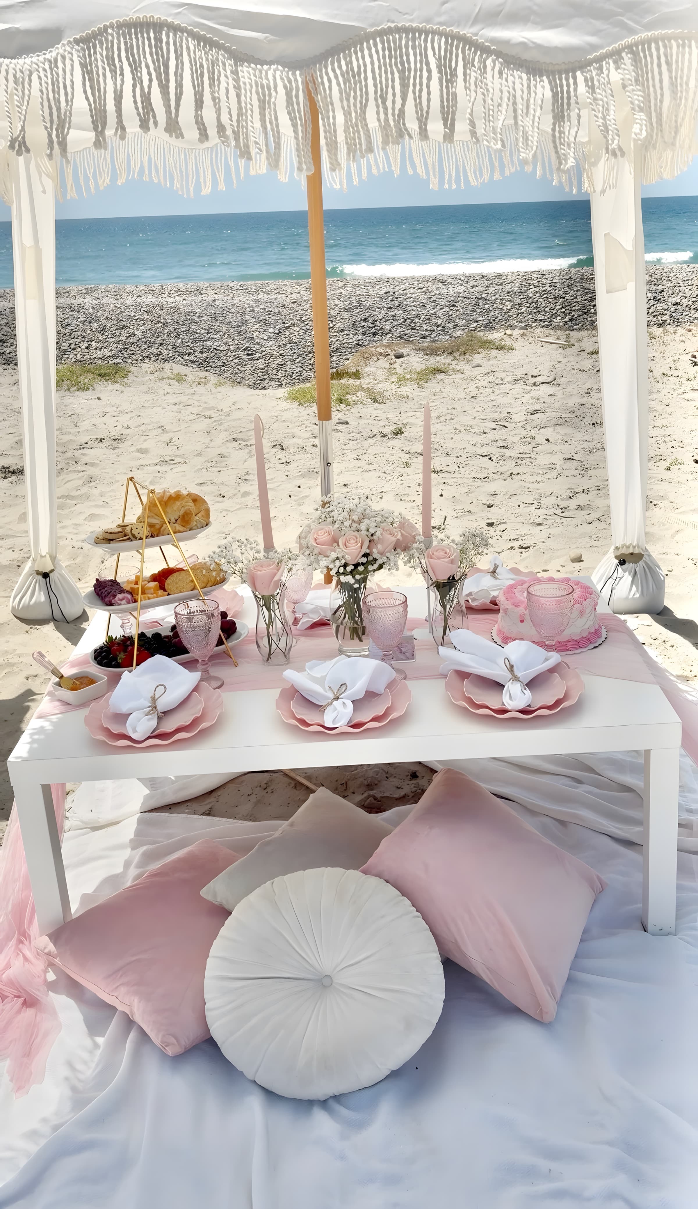 A blush beach picnic under a macramé-fringe umbrella with pink scalloped plates, roses and baby’s-breath, the ocean behind