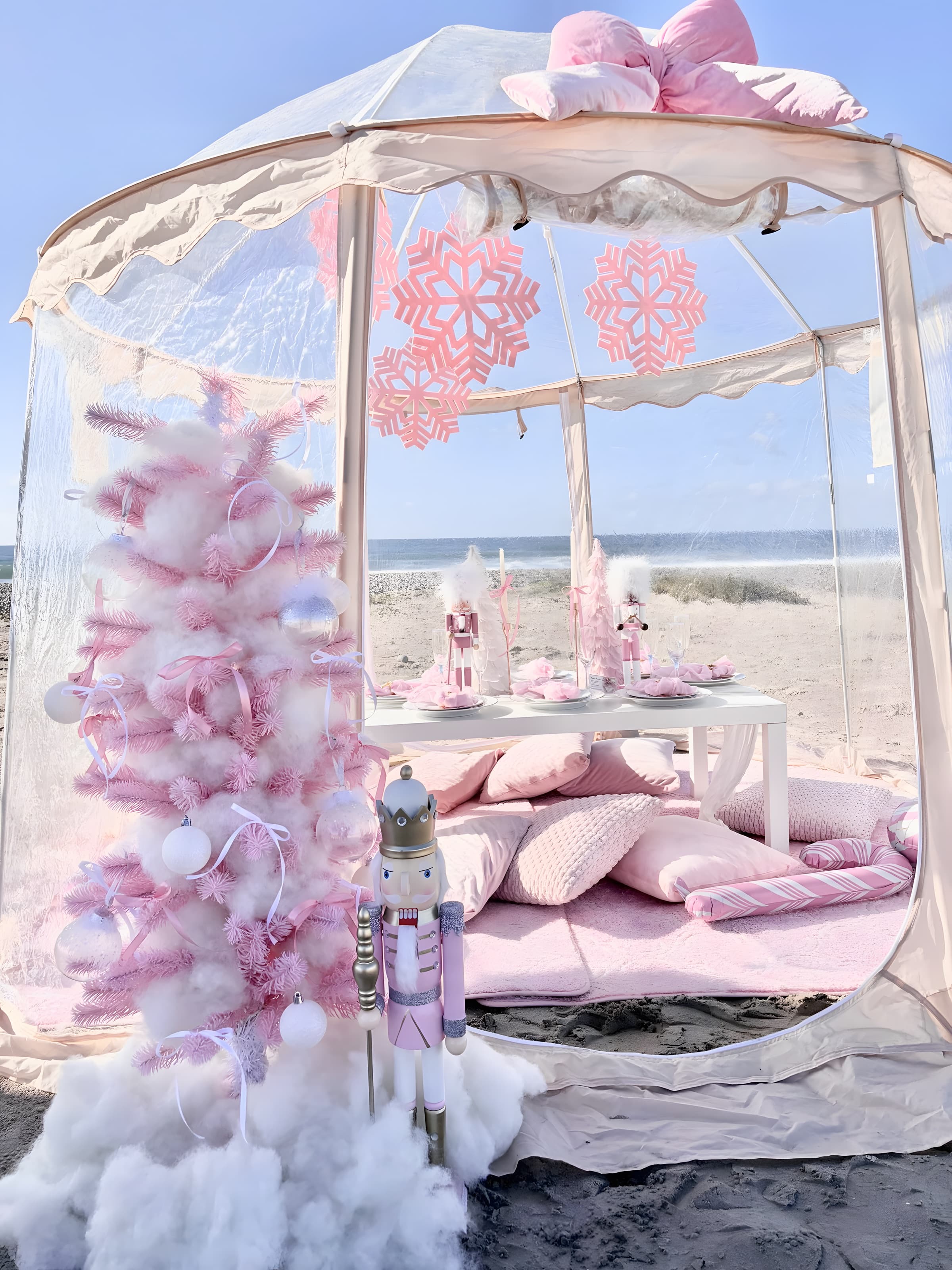 A clear bubble tent at the shoreline with a pink flocked tree and a set table, ocean beyond