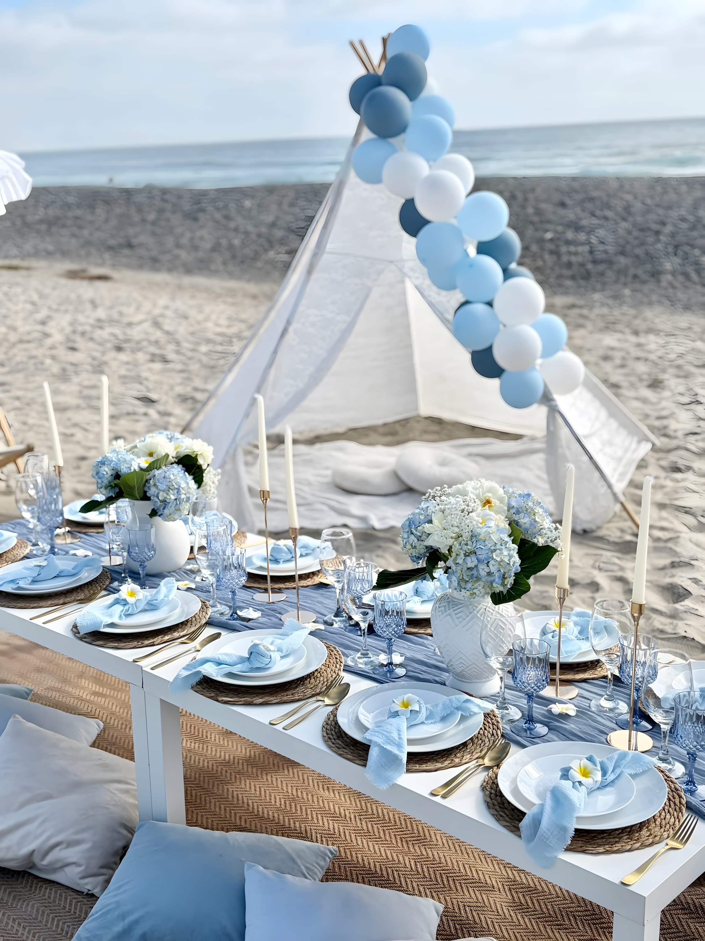A golden-hour dusty-blue setup with a lace teepee, balloon arch and hydrangea centerpieces, ocean behind