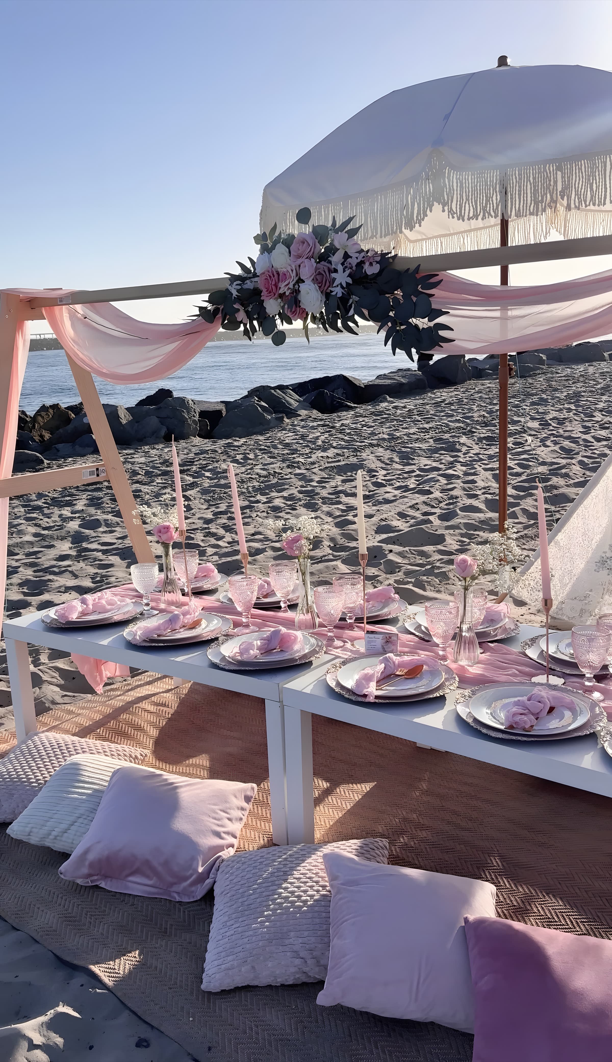A golden-hour blush beach picnic with a tasseled umbrella, a rose floral swag and pink goblets, the ocean behind