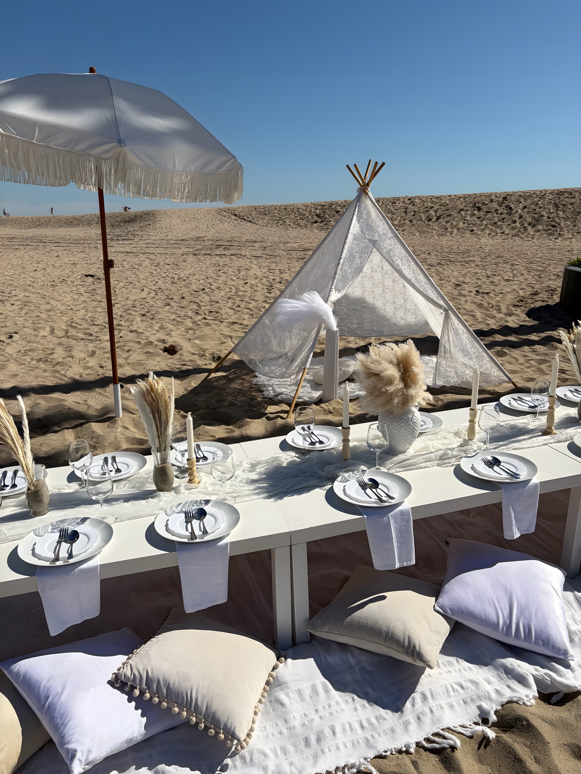 A neutral boho beach picnic with pampas centerpieces and pom-pom cushions toward a lace teepee and dune