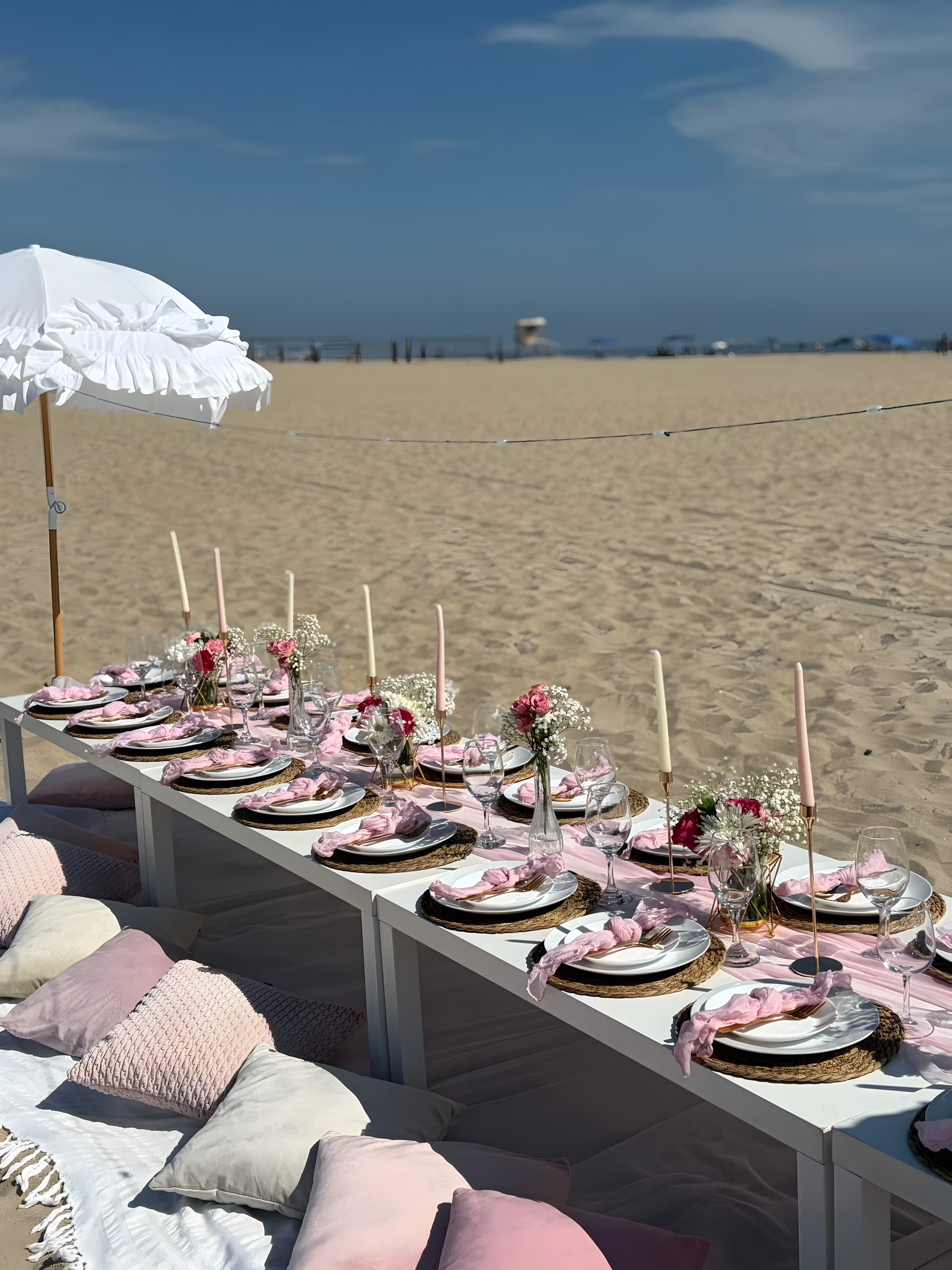 A blush beach tablescape down the table with a white ruffled umbrella, pink napkins and rose bud vases